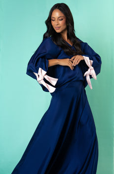Woman wearing a navy blue silk blouse with pale pink ribbon cuffs, paired with a matching flowing skirt, standing against a turquoise background.