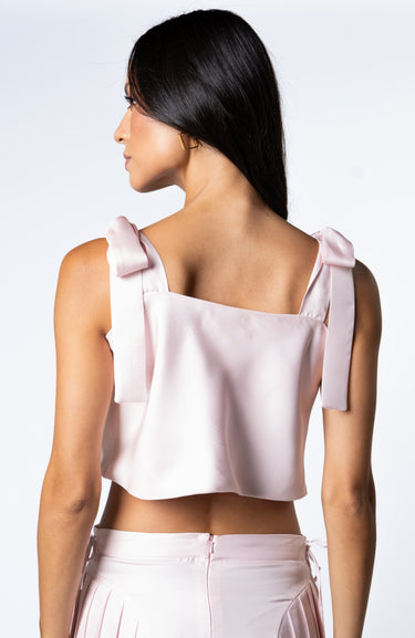 Back of a woman wearing a barely pink cropped silk tank with adjustable shoulder tie bows, paired with a matching high-waisted pleated silk skirt.