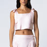 Woman wearing a barely pink cropped silk tank with adjustable shoulder tie bows, paired with a matching high-waisted pleated silk skirt.