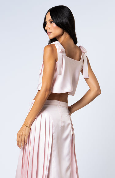Side view of a woman wearing a barely pink cropped silk tank with adjustable shoulder tie bows, paired with a matching high-waisted pleated silk skirt.